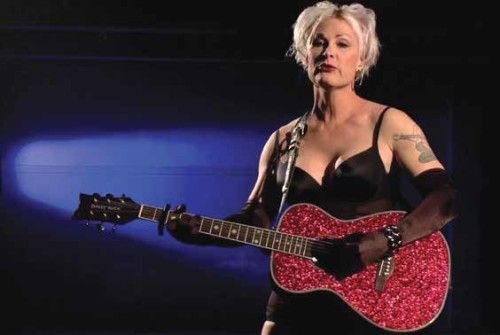 Shawna Virago stands in front of a microphone on a dark stage, strumming a sparkly-red guitar. She is wearing a short black sleeveless dress paired with black gloves and red lipstick. She has short platinum-white hair and a shoulder tattoo.