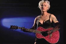 Shawna Virago stands in front of a microphone on a dark stage, strumming a sparkly-red guitar. She is wearing a short black sleeveless dress paired with black gloves and red lipstick. She has short platinum-white hair and a shoulder tattoo.