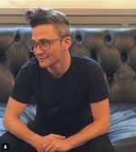 Photograph of Sam Feder seated on a couch looking off to the side with a slight smile. Sam is a white person with short hair and glasses. They are wearing a simple black t-shirt and are sitting in a relaxed leaning position with arms resting on their knees.