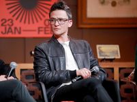 Sam Feder sitting on a panel listening attentively in a well-decorated space. They are seated holding a microphone, and other chairs are partially visible in the frame. Sam is a white person with short dark hair and glasses. They are wearing a leather jacket, a light shirt, and dark pants.