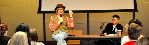 A wide photograph capturing both the audience and panel speakers, Kate Bornstein and Sam Feder. Kate Bornstein, a white person with an ear-length bob, brown hat and round glasses, sits on the panelist's table in an engaging pose while speaking into a microphone towards the audience. Sam Feder, a white person with short, dark coiffed hair and bold glasses, leans comfortably in a chair behind the table while listening to Kate.