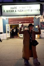 Friendly photograph of Kate Bornstein outdoors, standing proudly pointing with both hands to a cinema marquee above her that has her name and film promoted. Kate, a white person with a short light bob, round glasses and a statement hat. She is wearing a stylish long coat, a cross body bag and flared jeans. The picture was taken in the evening and is illuminated by the glowing marquee.