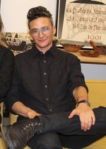 Cropped photo of Sam Feder seated, holding one leg crossed in their lap. Another person is partially visible on the left of the image. They picture is taken indoors in a room with artifacts in the background.