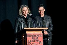 Amy Scholder and Sam Feder on a podium on stage. Amy Scholder, a white person with shoulder length light hair, is speaking into the microphone. She is wearing a leather jacket with a black hoodie underneath. Sam Feder, a white person with dark coiffed hair and glasses, stands beside her and smiles to the audience. They are wearing a leather jacket on top of a dark shirt.