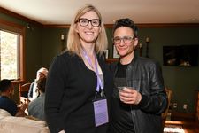 Sam Feder and Andrea James posing together for a photograph at a happy hour event taking place indoors in a living room. Sam is a white person with dark coiffed hair, bold glasses. They are wearing a leather jacket with a dark shirt underneath. Andrea James is a white person with shoulder length light hair and dark glasses. They are wearing dark clothing and a purple lanyard.