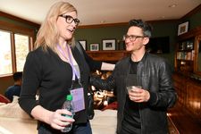 Candid photo of Sam Feder and Andrea James in a comfortable indoor living room, mid-conversation with an arm around each other. Sam is a white person with dark coiffed hair, bold glasses. They are wearing a leather jacket with a dark shirt underneath. Andrea James is a white person with shoulder length light hair and dark glasses. They are wearing dark clothing and a purple lanyard.