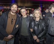 Sam Feder in linked arms with Amy Scholder and another person at a busy bar venue. Sam Feder is a white person with dark coiffed hair and bold glasses. They are wearing a leather jacket on top of a dark shirt and dark pants. Amy Scholder is a white person with wavy blonde shoulder-length hair, wearing a leather jacket. The other person is quite tall, white, with short dark hair and a beard. They wear a dark hoodie over a brown shirt, with a pair of glasses draped around their neck.