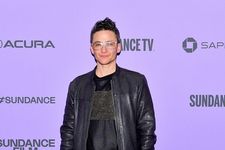 Sam Feder photographed on the Sundance red carpet against a press background with various sponsor logos printed. Sam Feder is a white person with dark coiffed hair, bold glasses, and dark clothing.