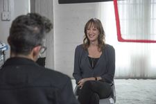 Behind the scenes moment of Jen Richards on set being interviewed by Sam Feder off screen. Jen is a white person with light brown shoulder length hair. Sam Feder, a white person with dark grey coiffed hair and glasses, can be seen from the back and out of focus. The set is a spacious and airy white room decorated with sheer curtains and a fluffy white rug.