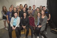 Behind the scenes photograph of 18 cast and crew members on set, sitting on apple boxes and standing. Camera gear and backdrops are visible in the background.