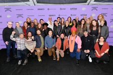 Large group photo of 30 cast on a festival red carpet. The colourful backdrop is sprinkled with festival and sponsor logos.