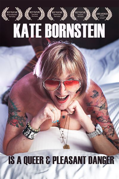 Kate Bornstein lies on her stomach on a bed with her chin propped on her knees and legs in the air. Kate is a white person, has a blond bob, and wears aviator sunglasses. Kate wears socks and is topless, showing off her tattoos and punk jewelry.
