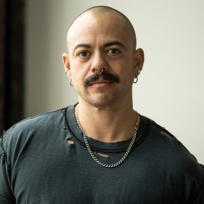 Headshot of Angelo Madsen Minax in his studio. He looks directly at the camera and wears a distressed grey t-shirt. He has light tan skin, a nearly shaven short buzzcut, thick mustache, and several small hoop piercings on the ears and a septum ring.