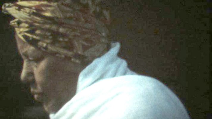 A close-up of a person's side profile as they look off-camera towards the ground. They have brown skin, and are wearing a patterned black-and-gold head wrap and a white outfit.