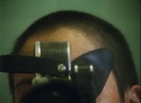 A close-up on someone's forehead and the top of their head. They are holding an object in front of their face. They have light skin and a buzzed head with dark brown hair. The object they are holding resembles a camera and is black and silver.