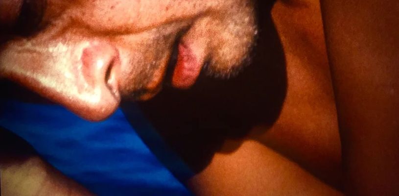 An extreme close-up of part of someone's face and chest in front of a blue background. They appear to be hunched over and folding into their body. They have light skin and are shirtless.
