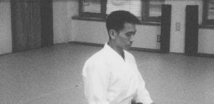 A black-and-white image of someone from the stomach up. They are sitting on a studio floor, wearing a white martial arts outfit. They have short dark hair and light skin.