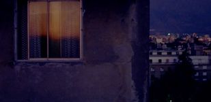 The edge of a building occupies the left two-thirds of the image. There is a visible window with light coming through but the shades are blocking any view into the window. The right two-thirds of the image features a skyline at night. The image has a blue tint over it due to the time of day.