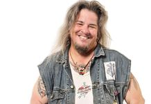 AAlec Butler has long sandy hair, a beard, light skin, and a big smile. They are wearing a denim vest with patches, a leather necklace with a pendant, and have some tattoos and piercings.