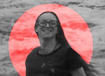 A black and white photo of Mirha-Soleil Ross. A red, slightly transparent circle frames her. The image is slightly blurry, but we can make out that Mirha-Soleil Ross has light skin, oval glasses, and long hair picked up by a breeze.