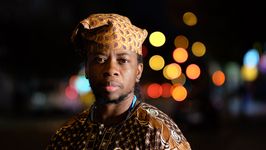 Seyi Adebanjo has an attentive expression, with the right side of their face well lit while the left is mostly in shadow. Seyi is a gender non-conforming Nigerian with a medium skin tone and short beard. They wear a loose headcovering and shirt with bold prints, as well as beaded necklaces and small earrings.