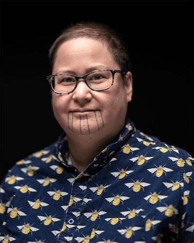 Theo Jean Cuthand wears glasses, has short hair, and is a light-skinned person of Plains Cree and Scots descent. He has facial tatoos on his chin of dots and vertical lines. He wears a smart button-up with a print of bees.