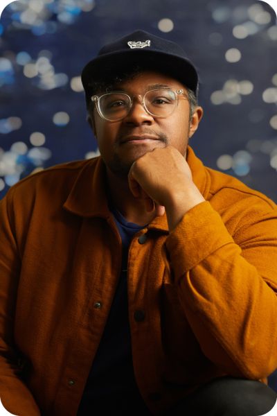 Brit Fryer looks pensive while resting his chin on his fist. Brit wears clear glasses, is a Black person with medium-skin tone, and has a slight mustache and short hair. He wears a baseball cap and a heavy button-up over a t-shirt.