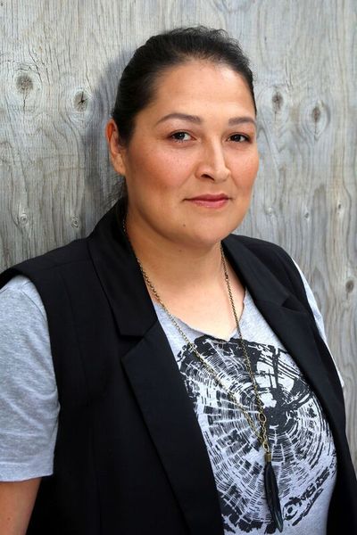 Cherish Violet Blood has dark hair styled into a ponytail, is a Blackfoot person, has light skin, and wears subtle makeup. Cherish wears a vest over a t-shirt with a silkscreeed tree ring crossection and a necklace with a feather pendant.