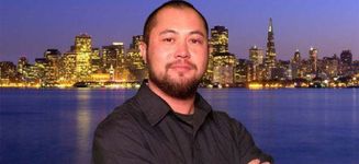 Christopher Lee looks at the camera with a neutral expression in front of a body of water and a nighttime city skyline. Christopher is an East Asian and white person, with a buzzcut, beard, and light skin. He wears a black button-up shirt.