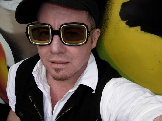 Selfie of Del Lagrace Volcano in front of a mural. Del has chunky glasses with dark lenses, light skin, short hair, and a slight goatee. They wear a ball cap, button-up shirt and a vest.