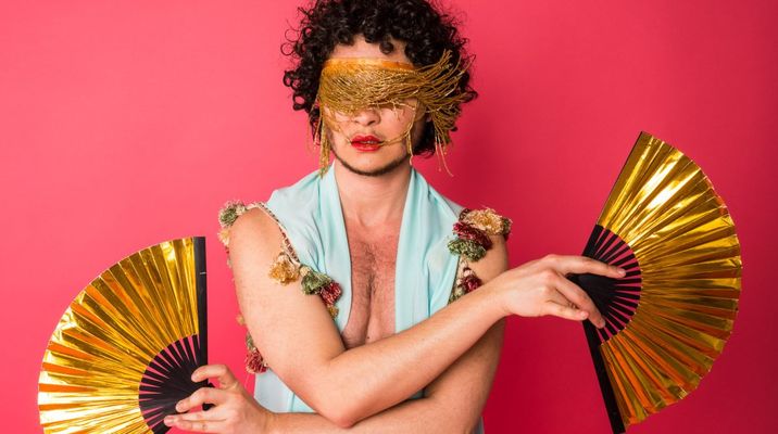 zavé martohardjono holds a gold folding fan in each hand. They wear a gold fringe headband which obscures their eyes. zavé is an Indonesian-American, with curly short hair, light skin, and short facial hair. They wear a vest with upholstry-style tassel and lipstick.