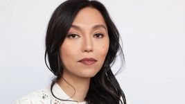 Isabel Sandoval has a neutral expression with slightly parted lips. Isabel is a Filipina person with long dark hair in loose waves and light skin. She wears a white lace sweater and subtle makeup, and stands in front of a monolithic off-white background.