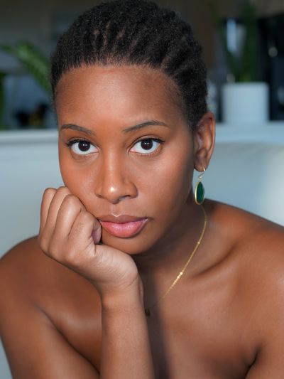 Nyala Moon, a Black Trans Woman, resting her face on her fist. She has light brown skin with brown eyes and short dark brown hair styled in braids. She is wearing green earrings lined in gold and a gold chain necklace. It is a shoulder up portrait focusing on her face.