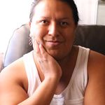 Cody L Makil is wearing a white tank top with light brown skin. He has one hand on his face and the other one is out of frame of the portrait. Cody has his nails a dark purple with his dark brown hair pulled back. He is wearing spiral shaped earrings.