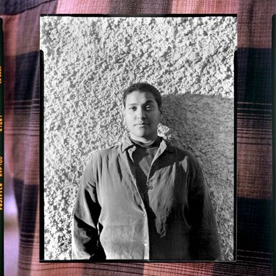 A picture within a picutre with a black and white portrait of shimby surrounded by a photo of plaid fabric. They are a Black person with short hair and a neutral expression. They wear a button-up over a turtleneck.