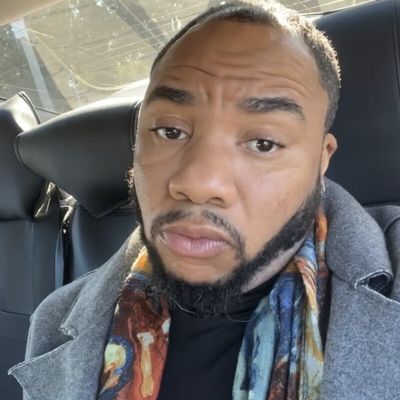 Kamar Porter in the back of a car with a neutral expression. Kamar is a light-skinned Black person with short dark hair and a beard. He wears a brightly patterned scarf and a peacoat.