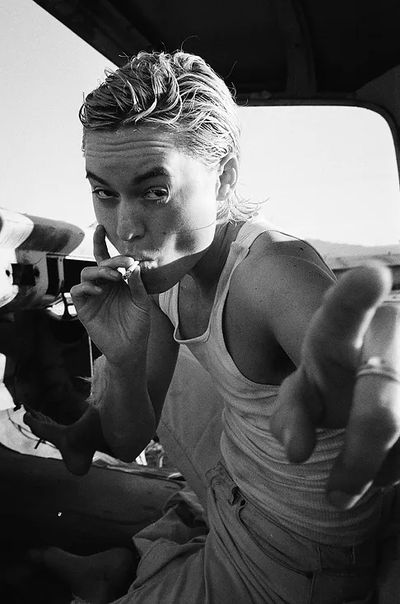 Black and white photo of Lío Mehiel in a car, smoking, and pointing at the camera. Lío is a Puerto Rican and Greek person with straight chin-length hair. They wear a muscle top and jeans.