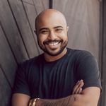 Lucah Rosenberg-Lee is a Black trans man with medium brown skin. He is shown from his chest up with his arms crossed and smiling. Lucah is wearing a short sleeve navy blue t-shirt. He has a tattoo on his right arm and a gold watch. Lucah is bald with dark facial hair and a chain tucked into his t-shirt. Lucah is in front of a dark brown wooden wall.