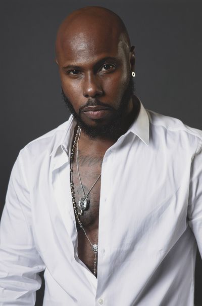 Marquise Vilsón with a focused expression. Marquise is a Black person with a shaved head, dark skin, facial hair, and tattoos on his chest. He wears a white shirt almost entirely unbuttoned with chains peeking out.