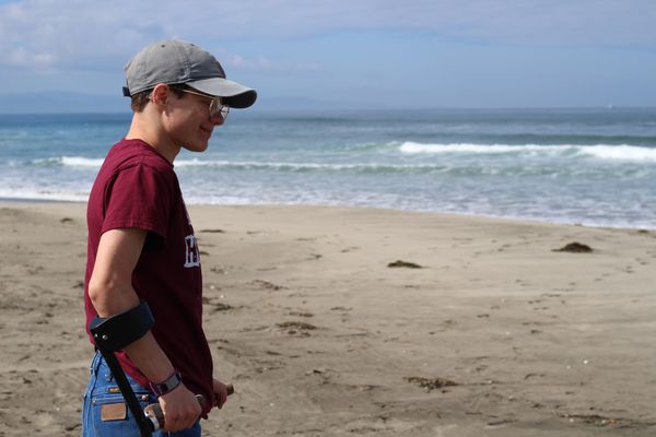 Marrok Sedgwick in profile on a beach. Marrok has forearm crutches, glasses, light skin, and short hair. He wears a baseball cap, t-shirt and jeans.