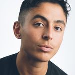Pidgeon Pagonis is Mexican and intersex with light brown skin in front of a white background. They are wearing a black t-shirt shown from the shoulders up with short dark brown hair with the sides faded. They have an industrial bar piercing on their ear.