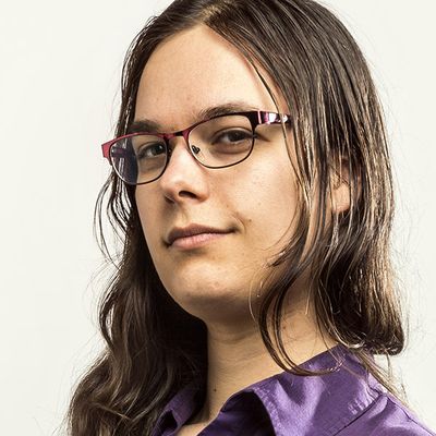 Tobi Hill-Meyer is an Indigenous Chicana trans woman with light tan skin in front of a white background. She is shown from the top of the shoulders up with a dark purple button up. She has long brown hair wearing small framed glasses.