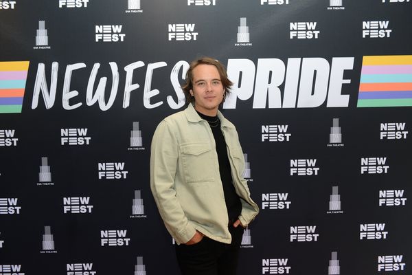Vuk Lungulov-Klotz is a Chilean-Serbian trans person. He has light skin standing in front of a banner that says, Newfest Pride. He is wearing a tan jacket over a black t-shirt with two gold chains and black pants. He has light blue eyes and short light brown hair that goes down to his neck and his hands tucked into his pockets.