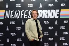 Vuk Lungulov-Klotz is a Chilean-Serbian trans person. He has light skin standing in front of a banner that says, Newfest Pride. He is wearing a tan jacket over a black t-shirt with two gold chains and black pants. He has light blue eyes and short light brown hair that goes down to his neck and his hands tucked into his pockets.