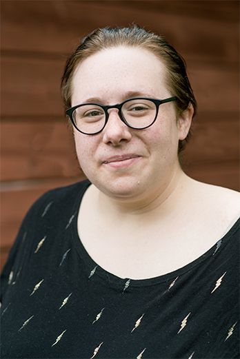 Jess Murwin is a nonbinary, mixed Indigenous settler with light skin. They have slicked back dark brown hair and are wearing black glasses. They are posing in front of a dark brown wooden background wearing a black t-shirt with small yellow lightning bolts across it.