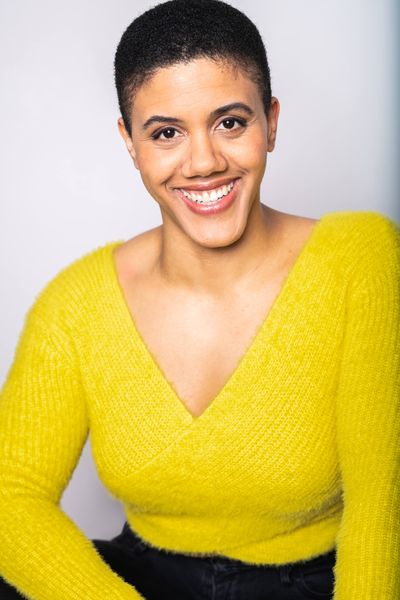 Amanda Cordner is Black and non-binary with light brown skin. They are posing in front of a white background in a yellow cut sweatshirt and black jeans. They have a buzzed head with black hair.