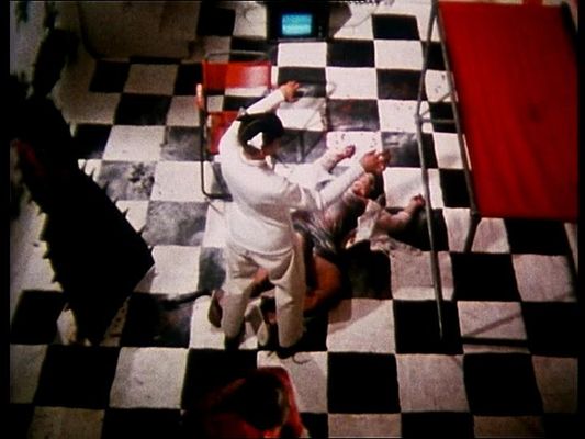 A fair skinned person dressed in a white uniform with black boots stands at the feet of a fair-skinned incapacitated person lying on the ground with minimal clothing. Both of their arms are raised near their heads and bent at the elbows, appearing to mirror one another. The setting is a graphic, high contrast room: large black and white tiles, a red chair and canopy, and a large black spikey standing object.
