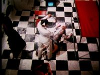 A fair skinned person dressed in a white uniform with black boots stands at the feet of a fair-skinned incapacitated person lying on the ground with minimal clothing. Both of their arms are raised near their heads and bent at the elbows, appearing to mirror one another. The setting is a graphic, high contrast room: large black and white tiles, a red chair and canopy, and a large black spikey standing object.