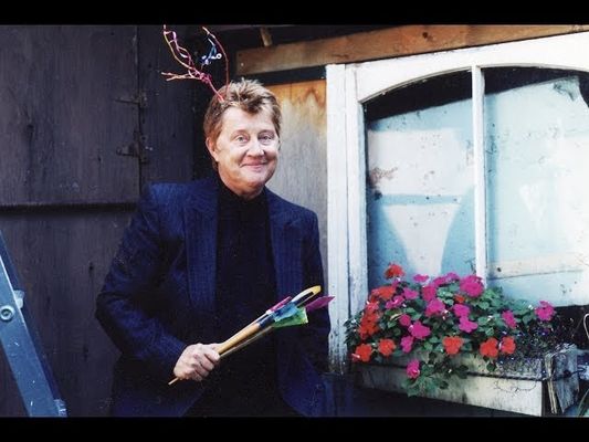 A person is in front of a wooden shed holding gardening tools. They are wearing a navy blue suit with a black turtleneck underneath with short dirty blonde hair. On the right hand side there is a window and a window sill flower box with purple and red flowers. They are smiling.
