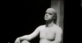 A fair skinned nude bald adult man sits on an empty black floor leaning back against a wooden beam. The person gazes down in a pensive manner. The still is in black and white.